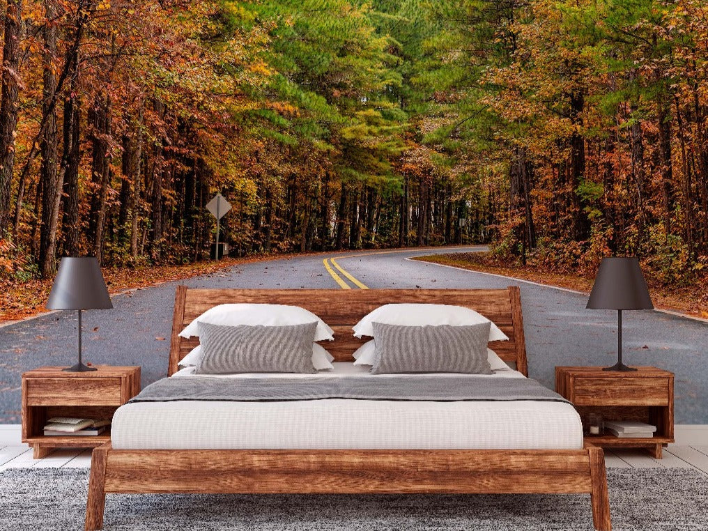 A cozy bedroom features a wooden bed with white and gray bedding, accompanied by two wooden nightstands with modern, dark lamps. The room's backdrop is a Decor2Go Wallpaper Mural of a winding path through a colorful forest called Autumn Road Wallpaper Mural, adding a touch of nature's tranquility to the space.