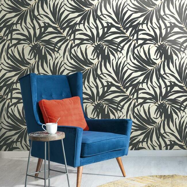 A stylish interior featuring a blue velvet armchair with an orange cushion beside a small round table. A white cup sits on the table, and the background boasts Black Bali Leaves Wallpaper by York Wallcoverings, adding nature-inspired charm to the sleek scene.