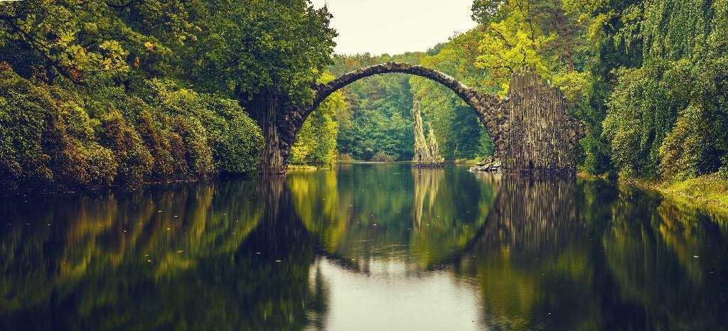 Ancient stone arch bridge spanning a calm river, surrounded by lush green trees, reflecting beautifully on the water surface in a Decor2Go Wallpaper Mural setting.