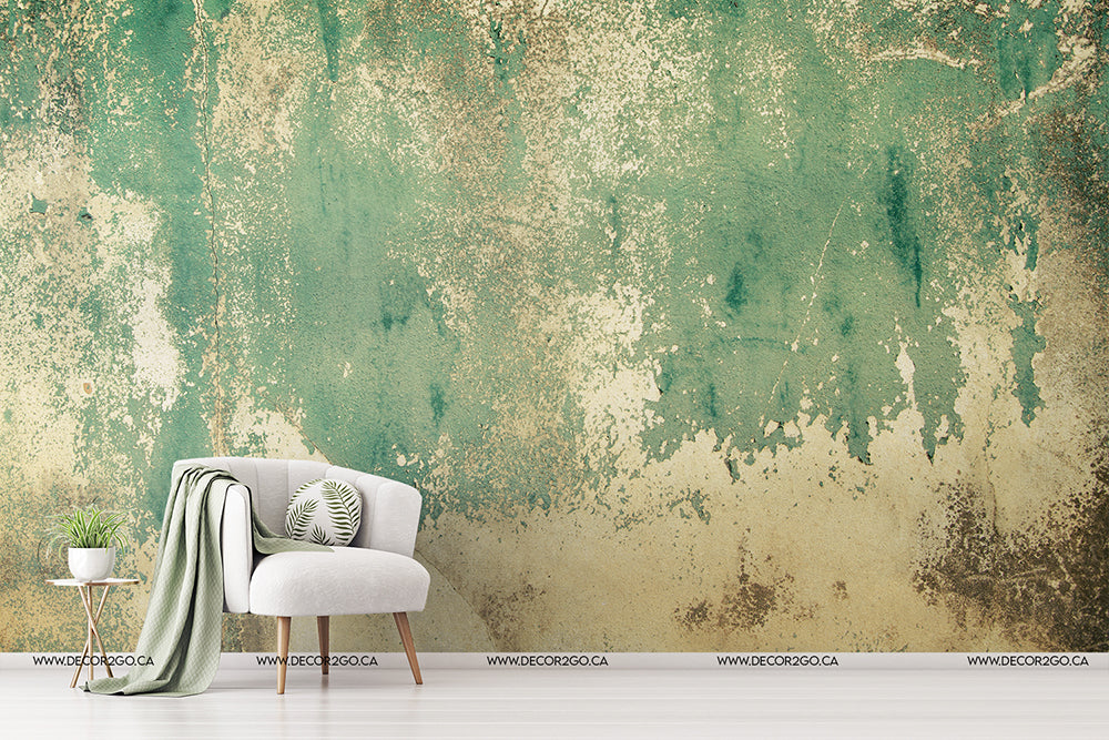 A white armchair with a green throw blanket and a cushion featuring a leafy pattern sits beside a small round table with a potted plant. The backdrop, reminiscent of the Rusti rust Weathered Concrete mural wallpaper from Decor2Go Wallpaper Mural, adds texture to the green and beige wall, creating an artistic atmosphere ideal for design studios.
