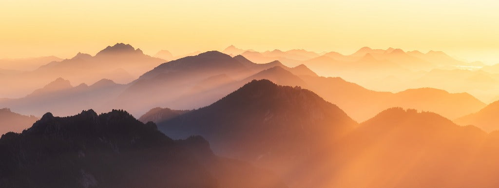 A panoramic view of layered mountain peaks under a warm, glowing Sunset on the Mountains Wallpaper Mural, with mist hovering between the ridges by Decor2Go Wallpaper Mural.