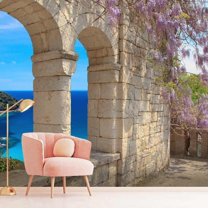 A pink velvet chair with a pillow sits next to a gold floor lamp, exuding rustic elegance. Behind them, the Decor2Go Wallpaper Mural's Tuscany Veranda View features a vibrant blue ocean and purple wisteria gracefully cascading down the walls through a stone arch.