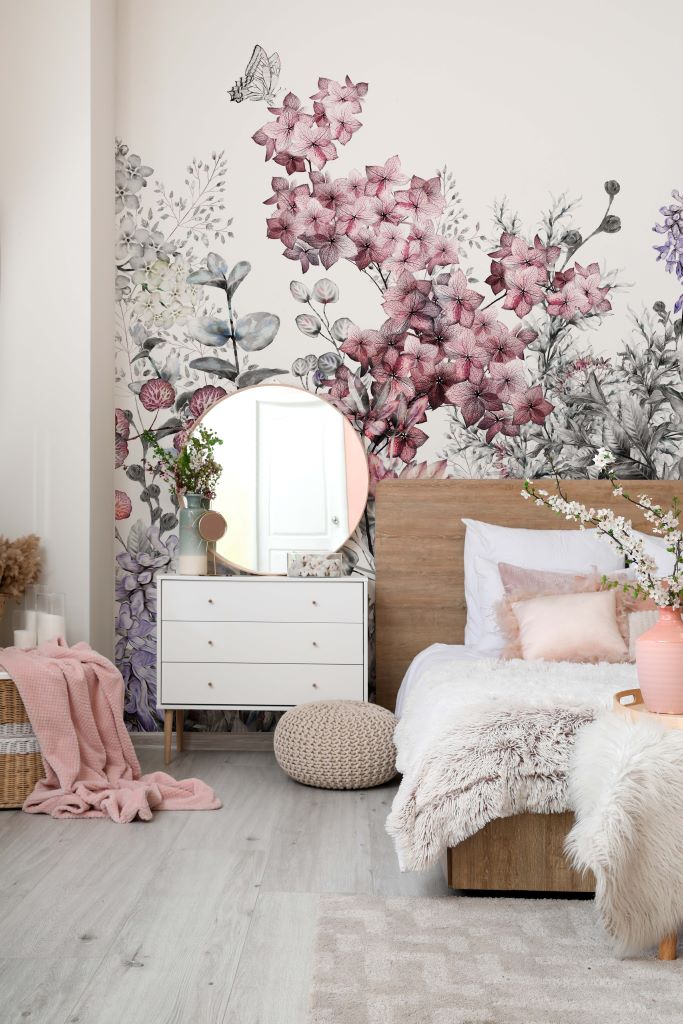 Bedroom - A cozy bedroom with a Wild Flower Wallpaper Mural feature wall, a wooden bed covered with soft white and furry textiles, a small white dresser with a round mirror, and decorative items like a vase and potted plants.