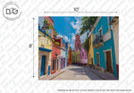 A vibrant colour palette street with houses in shades of blue, yellow, and red leading towards a pink church tower under a clear blue sky indicates the Decor2Go Wallpaper Mural Rainbow Road wallpaper design with dimensions labeled.