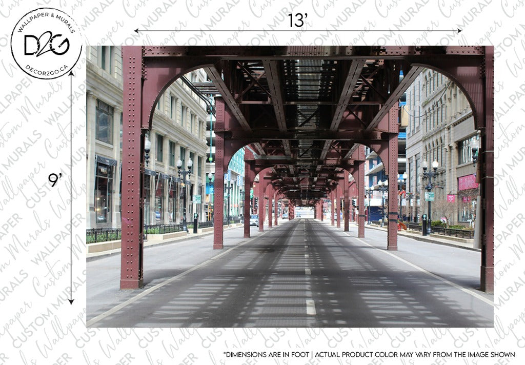 An image of a wide street beneath an urban railway track with red iron pillars. Buildings line both sides of the street, which is empty of cars and people. The dimensions of the vivid imagery are marked as 13 feet wide by 9 feet tall, featuring the Urban Railway Wallpaper Mural from Decor2Go Wallpaper Mural.