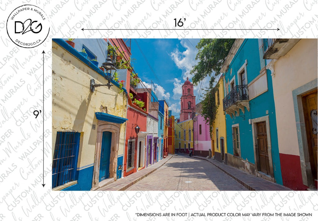 A vibrant street scene with colorful buildings lining a narrow road leading to a pink church tower under a bright blue sky, featuring custom sizing for the planned Decor2Go Wallpaper Mural.