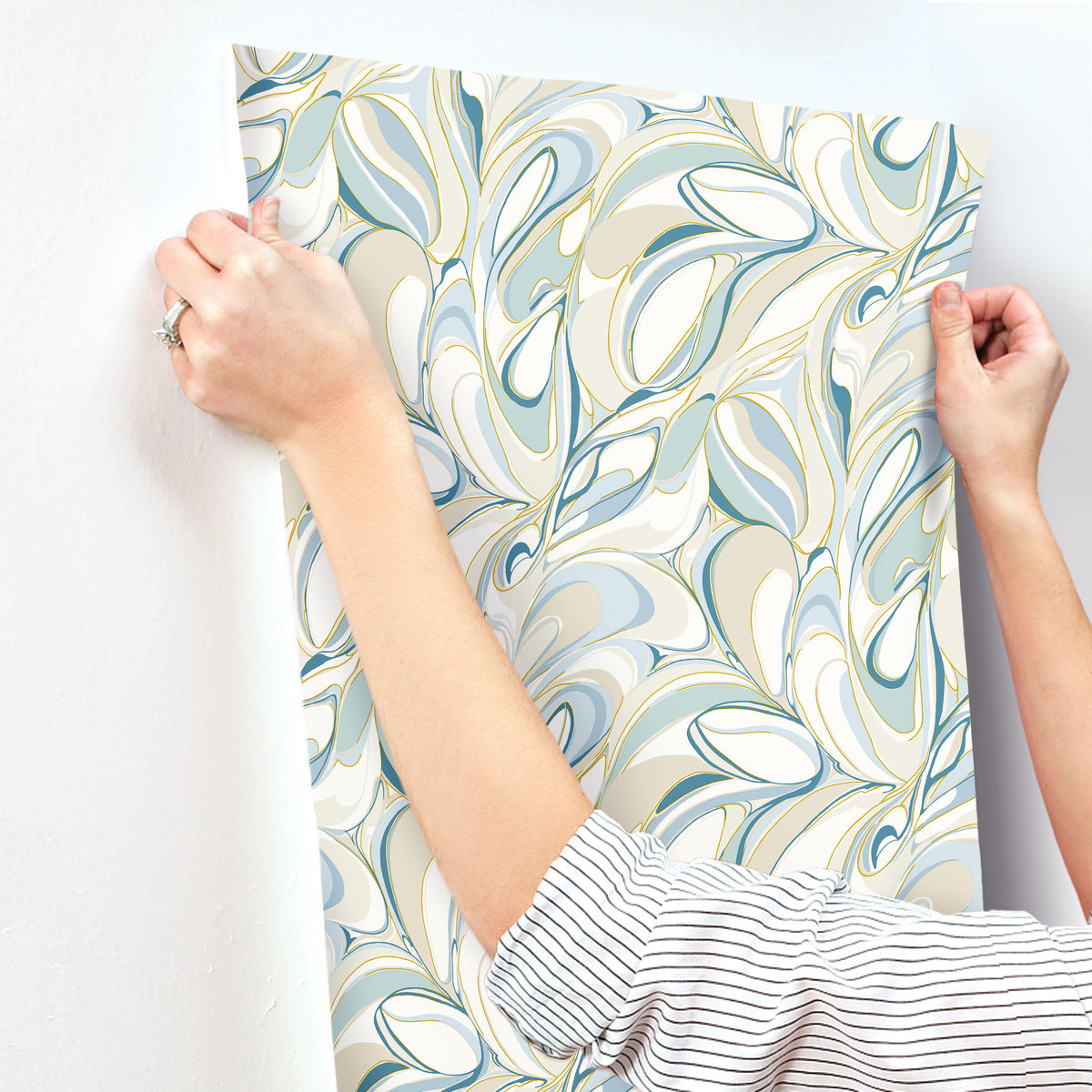 Only the hands and arms of a person in a striped shirt are visible as they apply York Wallcoverings Achate Blue Wallpaper (60 SqFt), featuring blue, green, and metallic gold marbled accents, to a white wall.
