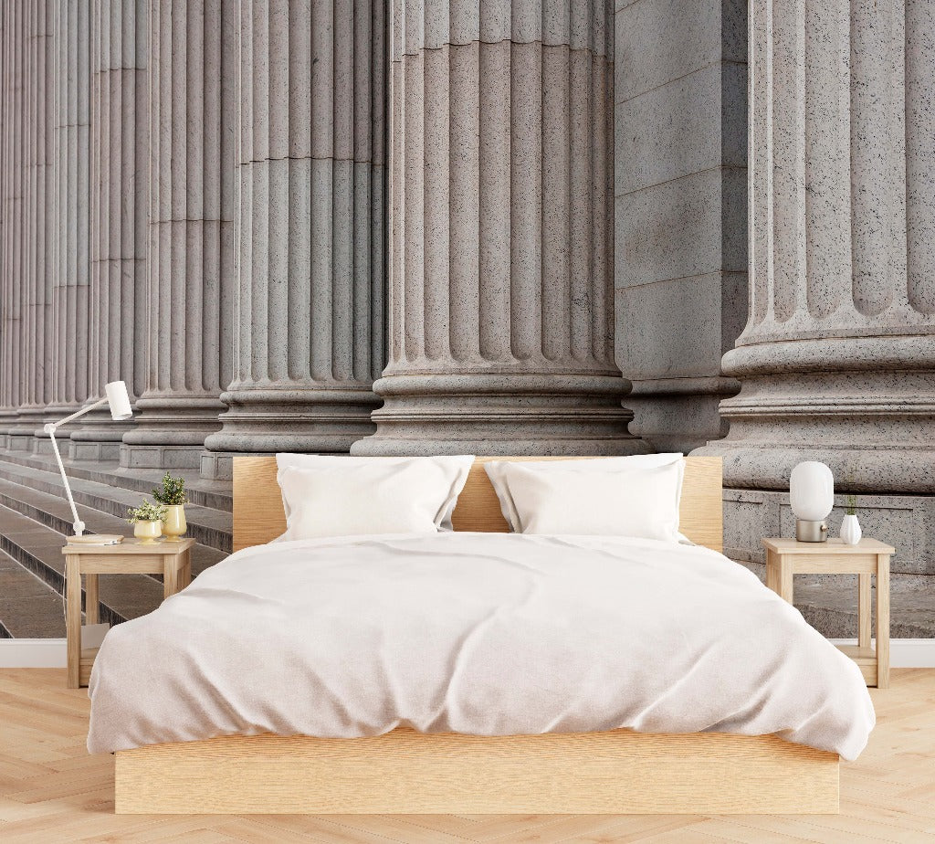 A modern bedroom with a large bed covered in a beige duvet, flanked by two wooden nightstands, against a backdrop of large, classical Ancient Pillars Wallpaper Mural inspired by Greek architecture from Decor2Go Wallpaper Mural.