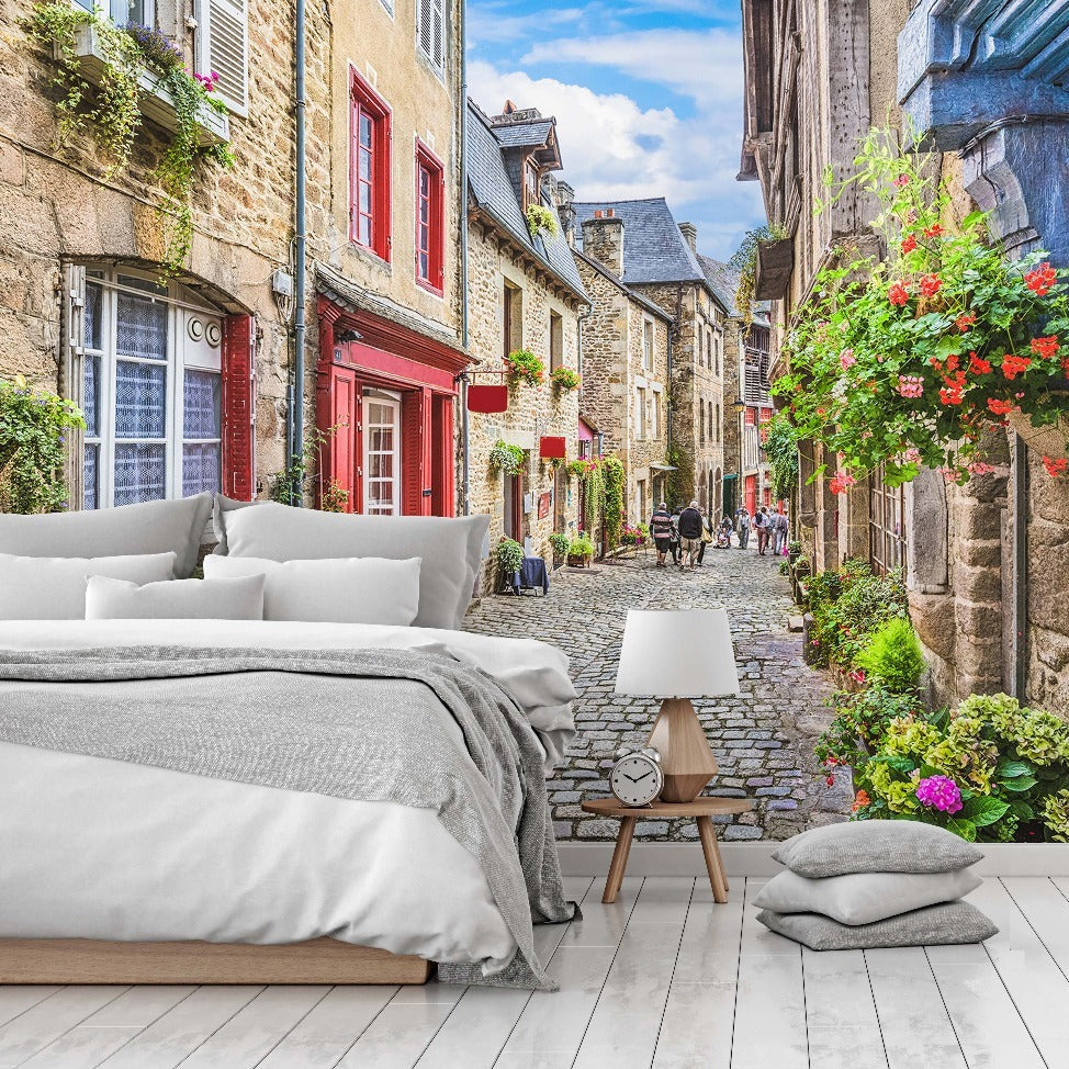 A digitally created scene combining a modern bedroom with an outdoor street view of a quaint European village. The bedroom features a neat bed and side table, seamlessly merging with a cobblestone street lined with colorful Antique European Alley Wallpaper Mural by Decor2Go Wallpaper Mural.