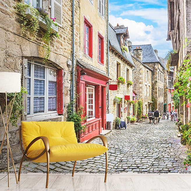 Cobblestone street in a quaint Decor2Go Wallpaper Mural with colorful houses, blooming flowers, and a modern yellow couch and lamp superimposed in the foreground. People are visible in the distance.