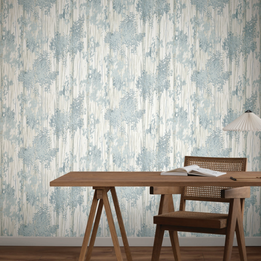 A wooden desk and a woven-back chair sit before York Wallcoverings’ Aspen Grove Blue Wallpaper (60 SqFt). An open book and modern white lamp rest on the desk, enhancing the wooden floor’s warmth.