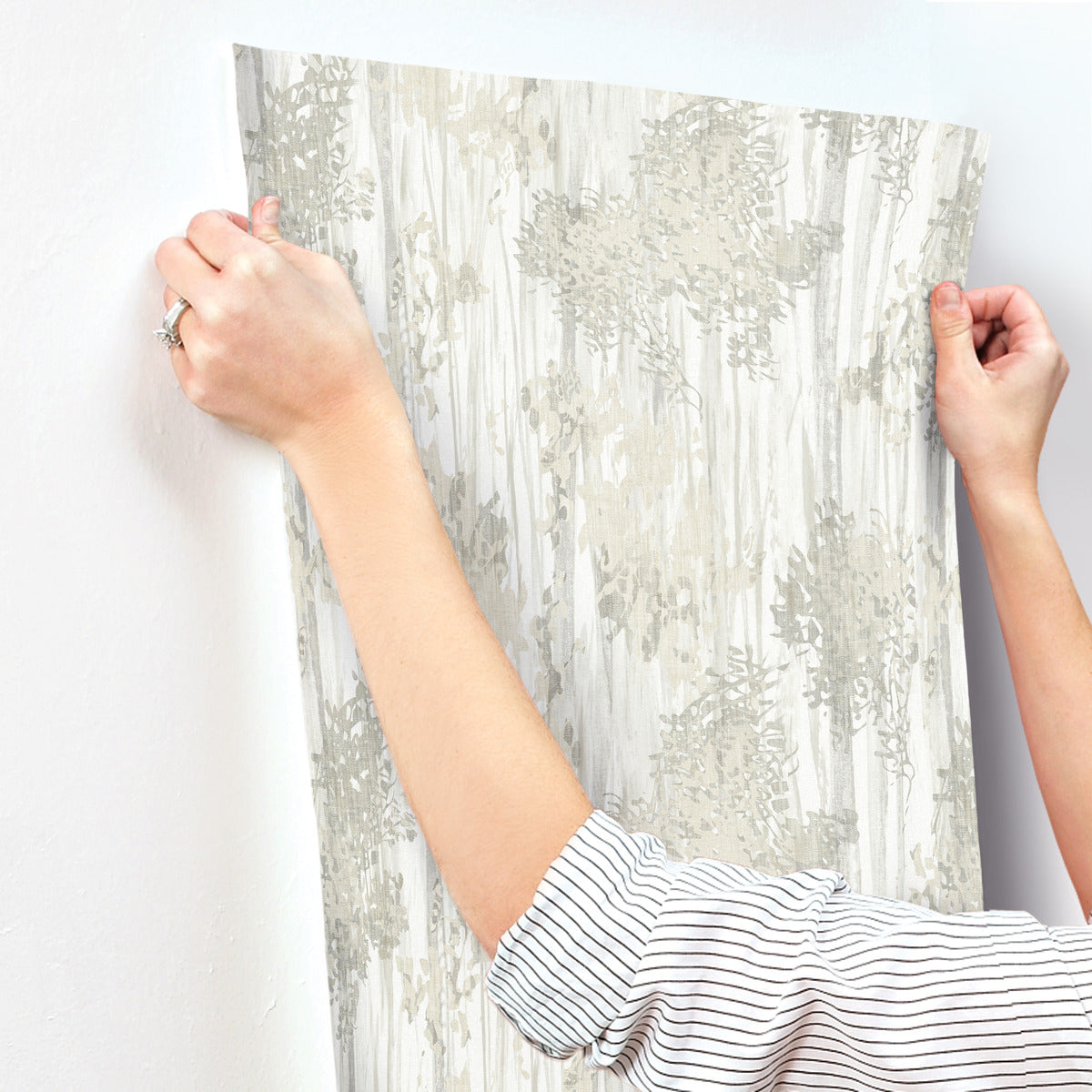 Wearing a striped shirt, a person uses both hands to install York Wallcoverings’ Aspen Grove Grey Wallpaper (60 SqFt) onto a white wall.