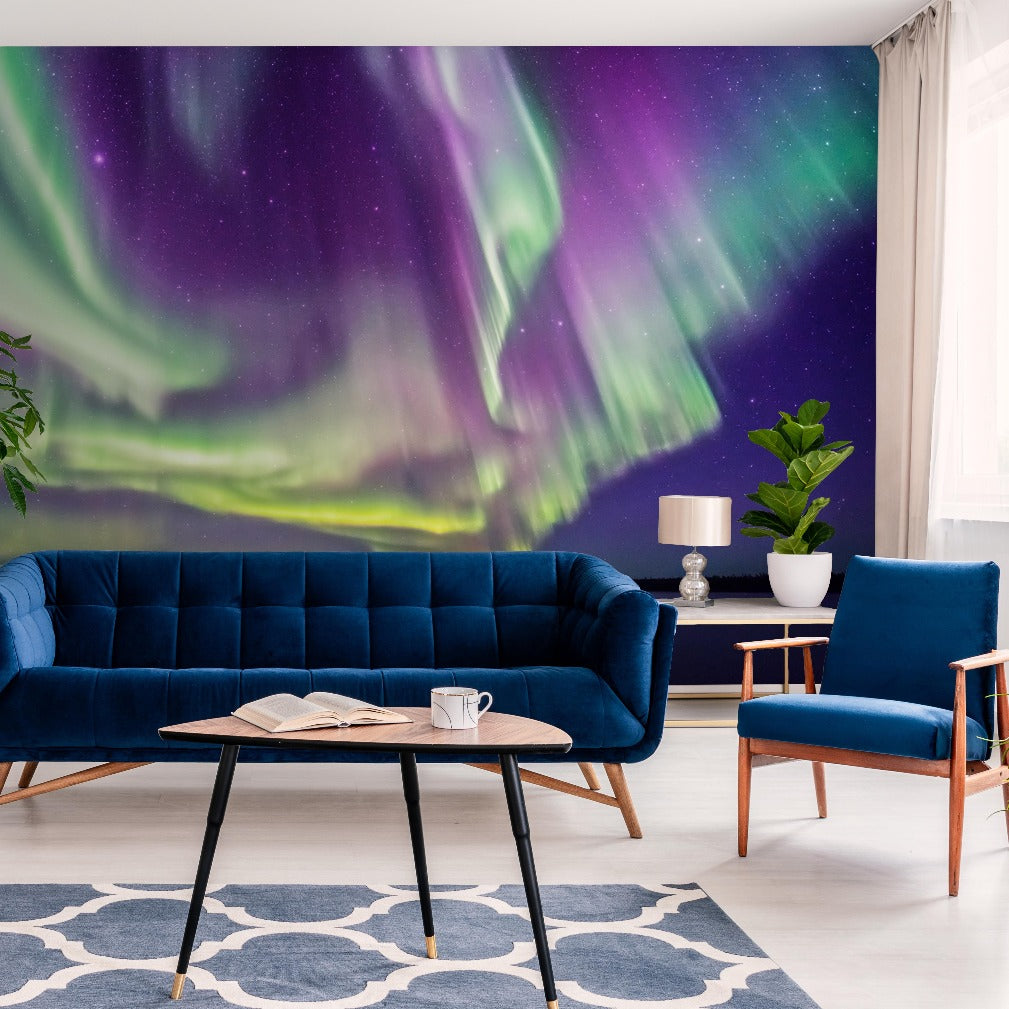 A modern living room with a dark blue velvet sofa, a matching armchair, and a black coffee table. The back wall features a Decor2Go Wallpaper Mural of the aurora borealis. Light decor and plants complete the scene.