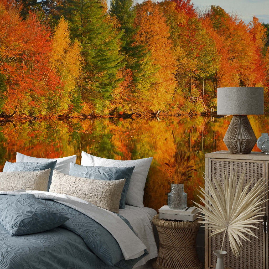A bedroom with a Fall Foliage Symphony Wallpaper Mural from Decor2Go depicting vibrant fall foliage reflecting in a lake, complemented by modern furnishings including a bed with pillows and a bedside table with a lamp and decorative items.
