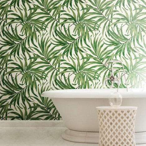A bathroom features a white bathtub and a wicker basket, with the wall behind adorned in York Wallcoverings' Green Bali Leaves Wallpaper (60 SqFt) creating a tropical atmosphere. The light-colored flooring enhances the serene, nature-inspired theme.
