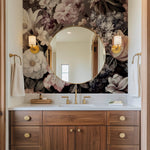 A bathroom interior featuring the Decor2Go Blooming Meadow Wallpaper Mural as a backdrop to a large round mirror. The wooden vanity is adorned with brass fixtures, a white countertop, and towels gracefully hanging on each side. Warm lighting from wall sconces enhances the decor's elegance.