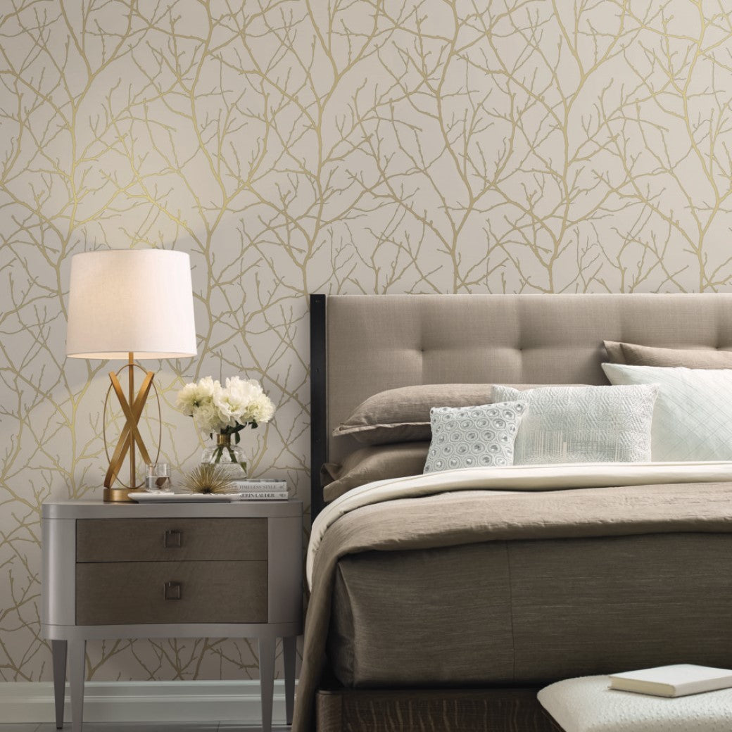 A bedroom with a tufted headboard and beige bedding is enhanced by metallic accents. Beside the bed is a nightstand with a lamp, books, and flowers. The room features York Wallcoverings' Beige & Gold Trees Silhouette Wallpaper, adding a subtle tree branch pattern.