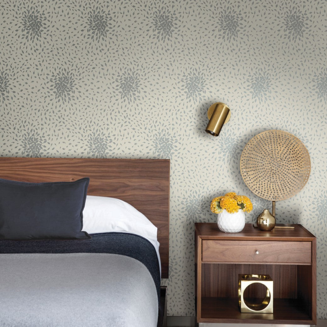 In a modern bedroom with a contemporary look, a wooden bed features gray and white bedding, accompanied by a nightstand holding yellow flowers and a rattan lamp. A gold wall sconce complements the York Wallcoverings Beige & Silver Petite Leaves Wallpaper for added elegance.