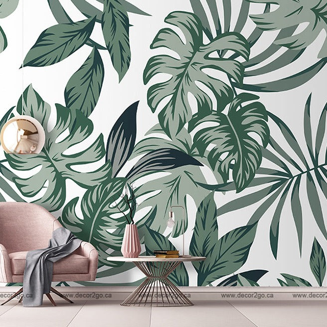 A stylish room with a Big Green Leaves Wallpaper Mural from Decor2Go, featuring a pink armchair, a small round table with books, a glass, and a bottle, and a wall-mounted round mirror.