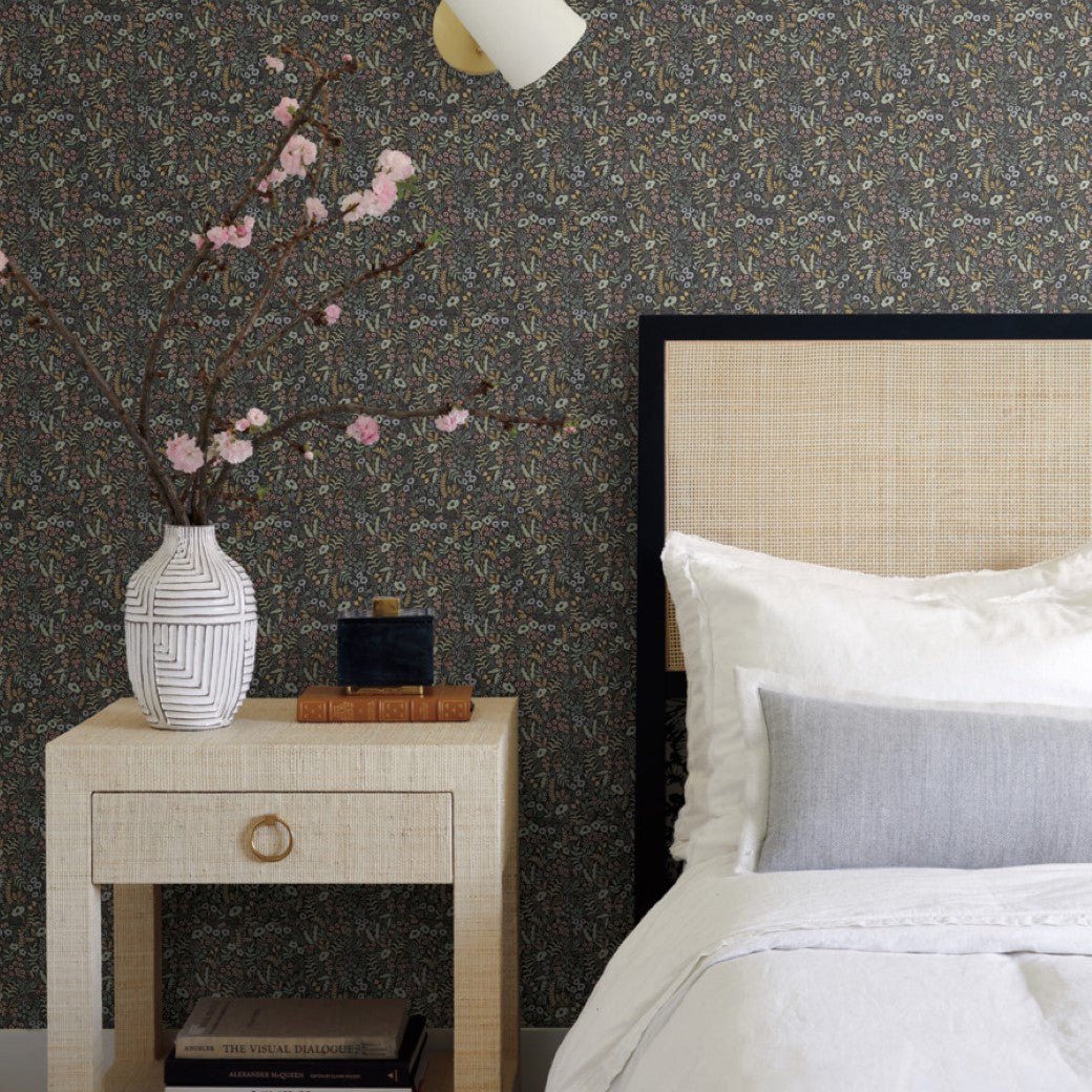 In this serene bedroom scene, a beige bed is adorned with white and gray pillows. The nightstand with botanical designs holds a vase of blossoming branches and stacked books. The wall features York's Black Tapestry Wallpaper topped by a modern light fixture above.