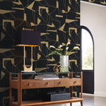 A modern interior showcases a wooden console table with two drawers. A black lamp with a gold base sits atop, next to a clear vase of white flowers and books. The wall features York Wallcoverings' Black & Gold Abstract Geo Wallpaper, and an arched window completes the scene.