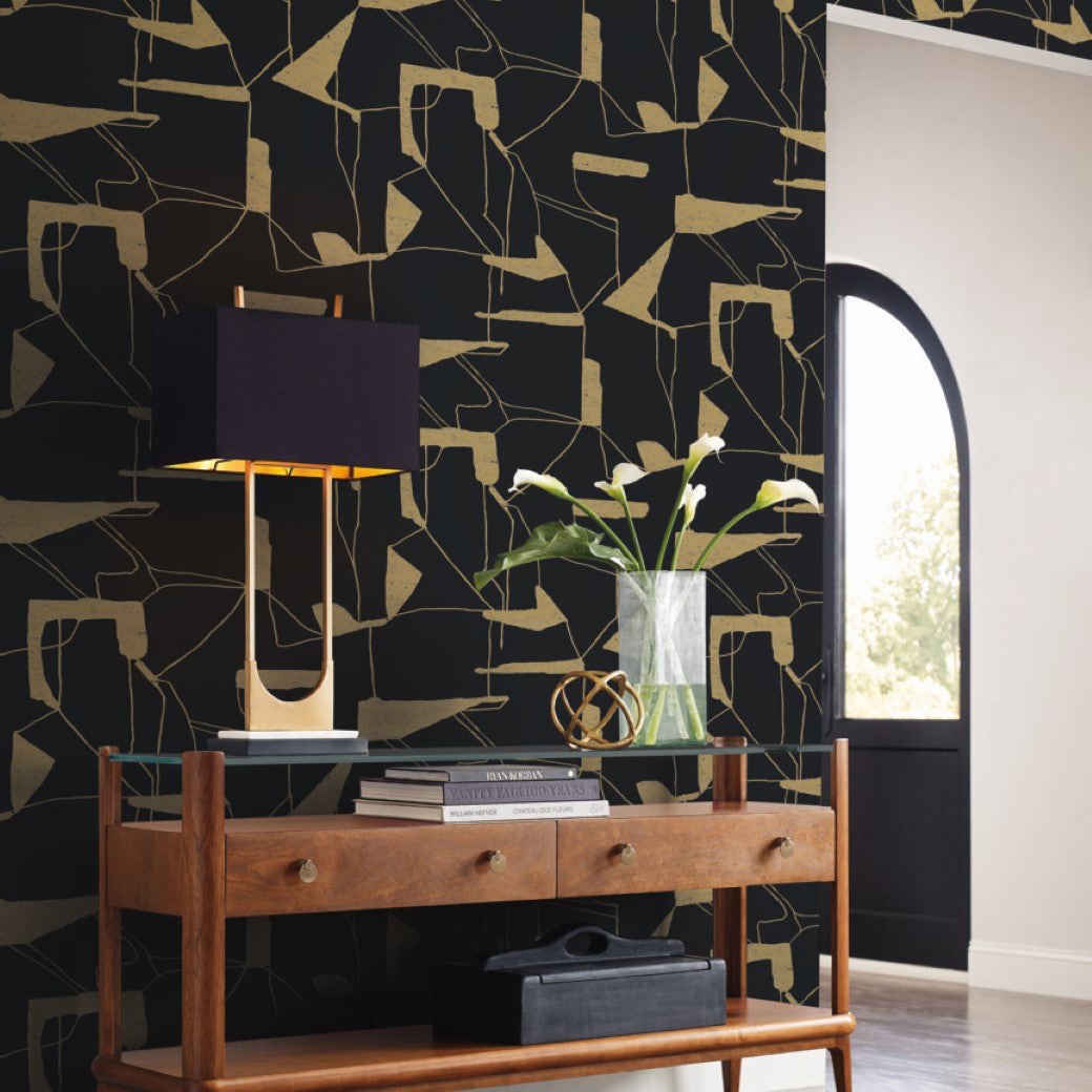 A modern interior showcases a wooden console table with two drawers. A black lamp with a gold base sits atop, next to a clear vase of white flowers and books. The wall features York Wallcoverings' Black & Gold Abstract Geo Wallpaper, and an arched window completes the scene.