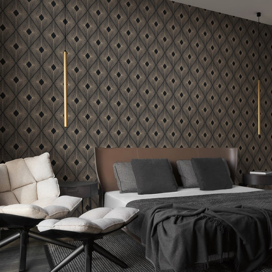 A modern bedroom features York Wallcoverings' Black & Gold Harlowe Wallpaper with geometric design, a large bed with dark pillows, gray blanket, two cushioned chairs in the foreground, and a vertical pendant light from the Modern Metals Collection adding a contemporary touch.