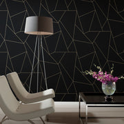Living Room - A modern with two white chairs and a tall floor lamp features the York Wallcoverings Black & Gold Nazca Wallpaper, showcasing geometric metallic gold lines on a black background. A black table with a vase of purple orchids adds color to the minimalist decor