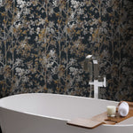 A modern bathroom scene features a white bathtub with a wooden tray holding a candle and rolled towel. The background is enhanced by York Wallcoverings' Black & Multi Shimmering Foliage Wallpaper, adding metallic accents. A sleek silver faucet graces the edge, enhancing the contemporary elegance.