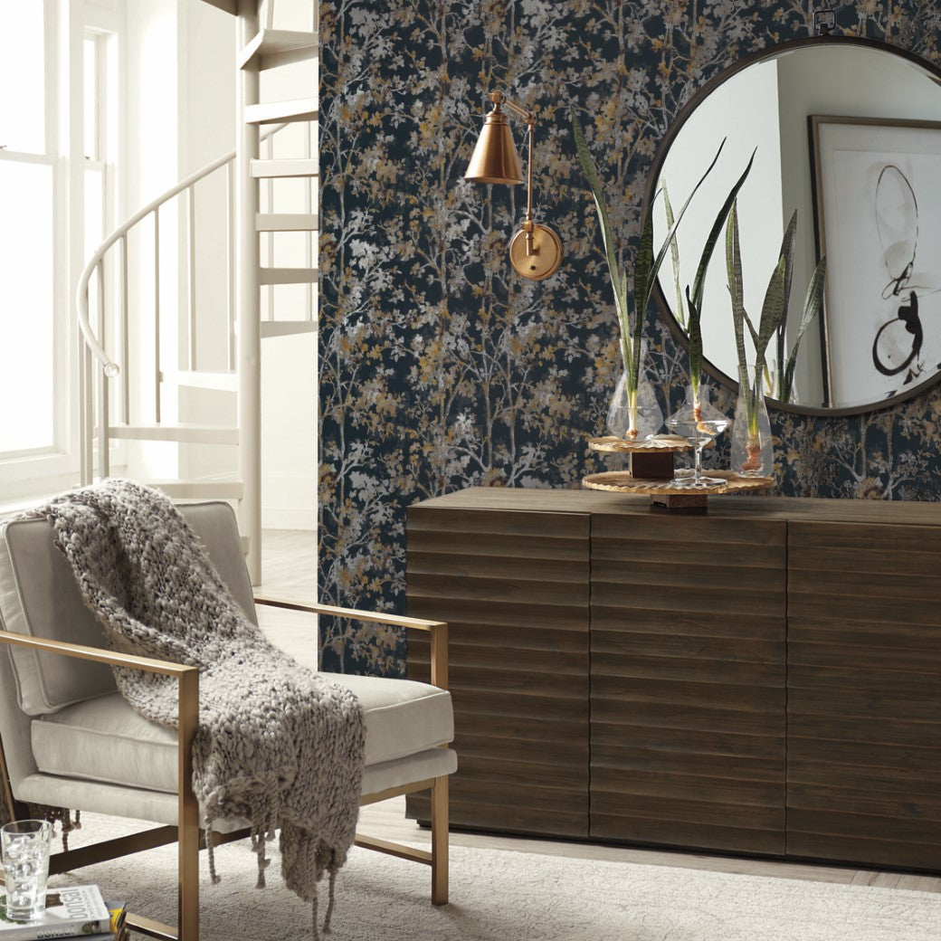 A cozy living room showcases a beige armchair with a chunky knit throw, a wooden sideboard, a round mirror, and an ornamental plant. The backdrop features York Wallcoverings' Black & Multi Shimmering Foliage Wallpaper, adding depth and elegance. A spiral staircase is in the background.