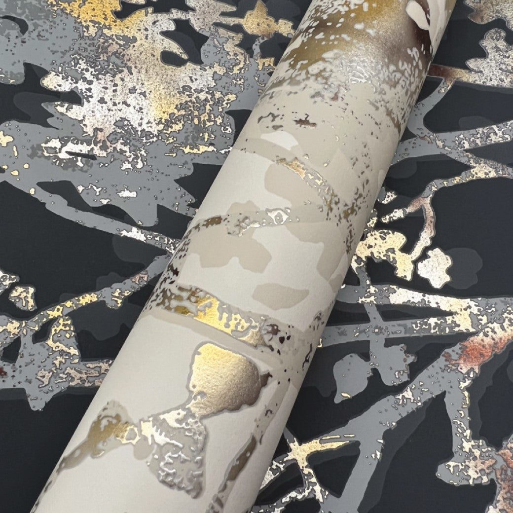 A roll of Black & Multi Shimmering Foliage Wallpaper by York Wallcoverings lies on a surface with gold, silver, and gray abstract metallic designs resembling organic shapes that shimmer in the light.