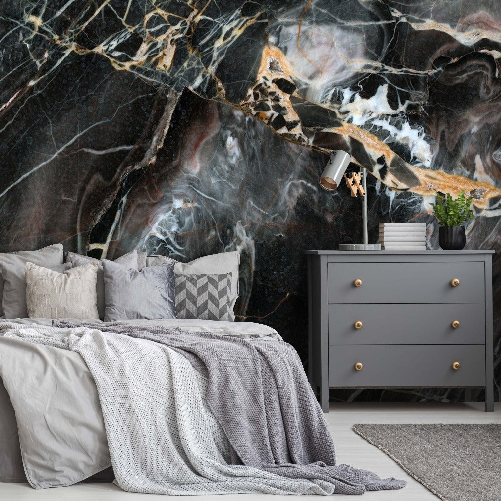 A modern bedroom featuring a large bed with gray bedding in front of a dramatic Decor2Go Wallpaper Mural black and gold marble wall. A gray dresser, small black side table, and houseplants complement the luxurious decor.