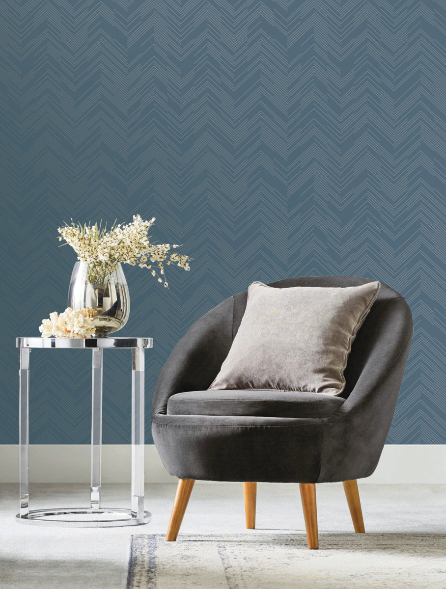 A cozy, modern living space features a dark gray armchair with a light gray pillow. A clear glass side table from the Modern Metals Collection holds a vase of white flowers, while York Wallcoverings' Blue & Silver Polished Chevron Wallpaper with an intricate geometric design enhances the background.