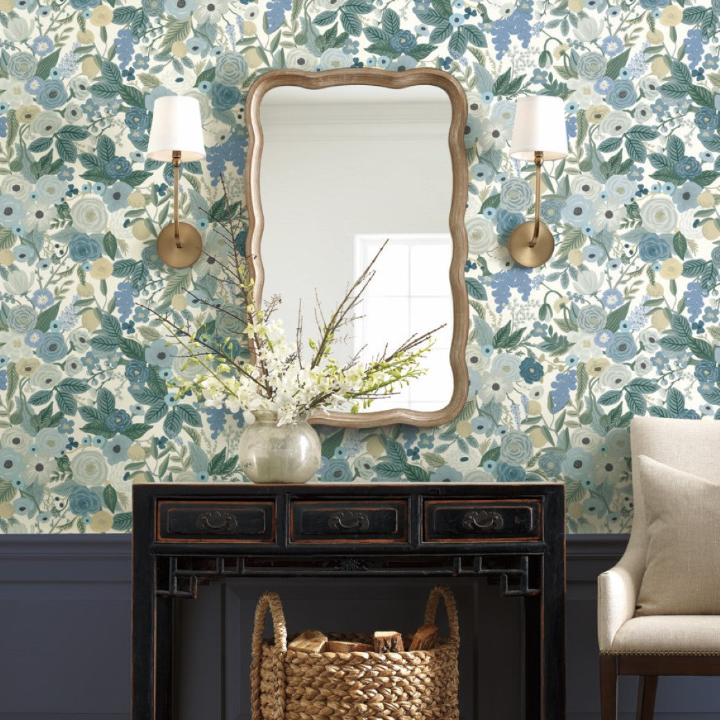 A wooden table with a wicker basket and white floral vase sits beneath a wavy-framed mirror. Two sconces flank the mirror on York's Blues Garden Party Wallpaper, featuring bold florals. A beige cushion is partially visible on the right.