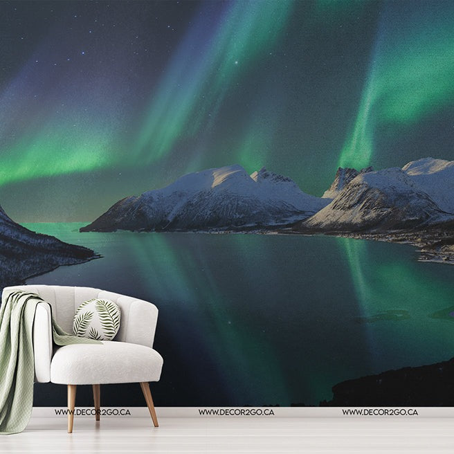 A modern interior with a white chair and a green throw blanket in front of a large wall mural displaying the Borealis Skies Wallpaper Mural by Decor2Go Wallpaper Mural over a serene mountain lake.
