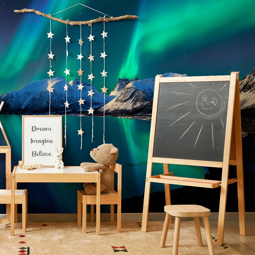 A cozy children’s room corner with a rustic theme, featuring a wooden easel with a chalk drawing, a teddy bear on a chair, and a Borealis Skies Wallpaper Mural over mountains from Decor2Go Wallpaper Mural.