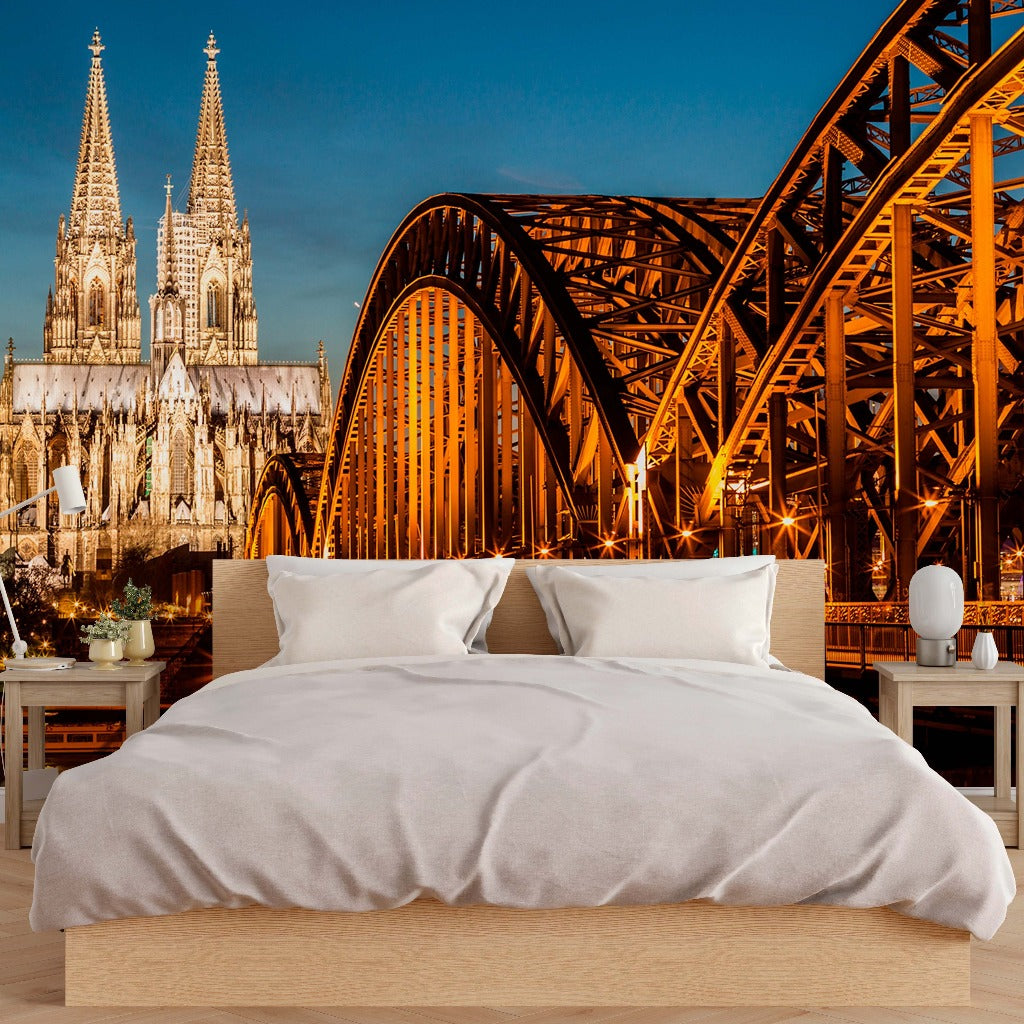 A creative bedroom setup with a wall-sized Bridge and Cathedral View Wallpaper Mural from Decor2Go Wallpaper Mural depicting the cologne cathedral and hohenzollern bridge at night, featuring a bed with white bedding in the foreground, designed to evoke.