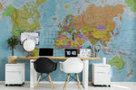 A modern office space with a large Decor2Go Wallpaper Mural of Bright World Map on the wall, featuring a wooden desk with a computer, two black chairs, and a small potted plant.