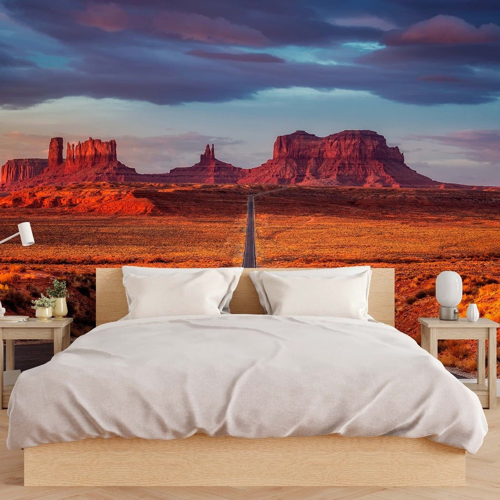 A modern bedroom with a large bed, nightstands, and lamps, with a wall-sized Decor2Go Wallpaper Mural displaying a vivid desert landscape featuring monumental red rock formations under a dramatic sky.