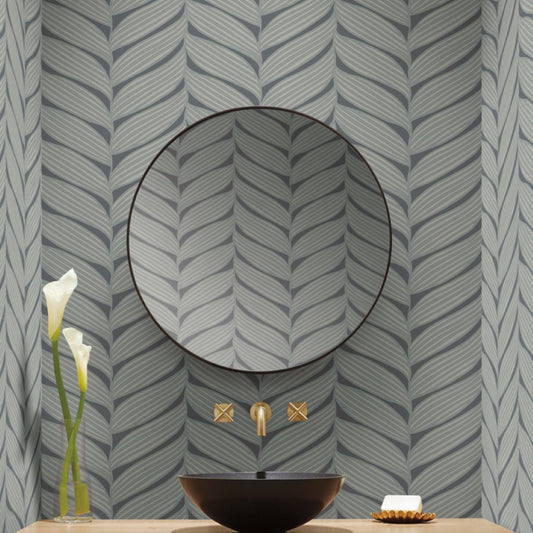 A modern bathroom features a large round mirror, an angled gold faucet, and a black bowl-shaped sink. Two calla lilies are in a vase on the left, with a soap dish on the right. The wall showcases York Wallcoverings' Charcoal & Silver Luminous Leaves Wallpaper.