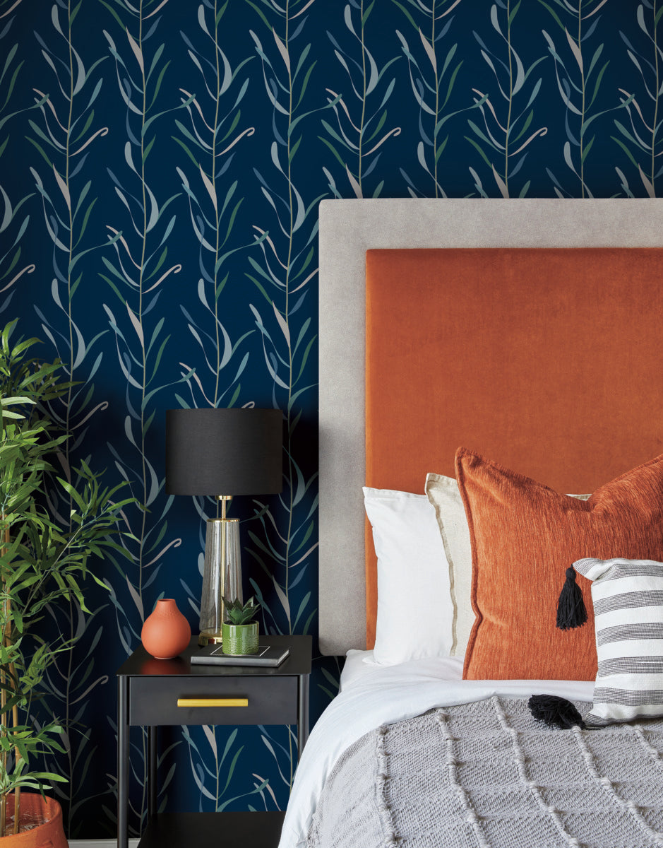 A cozy bedroom features a bed with an orange headboard and white, orange, and striped pillows. The wall behind showcases a whimsical design with dark blue Chloe Vine Indigo Wallpaper Blue, Green (60 Sq.Ft.) from York Wallcoverings. A black bedside table holds a black lamp with a gold base and various decor items, including a small plant.