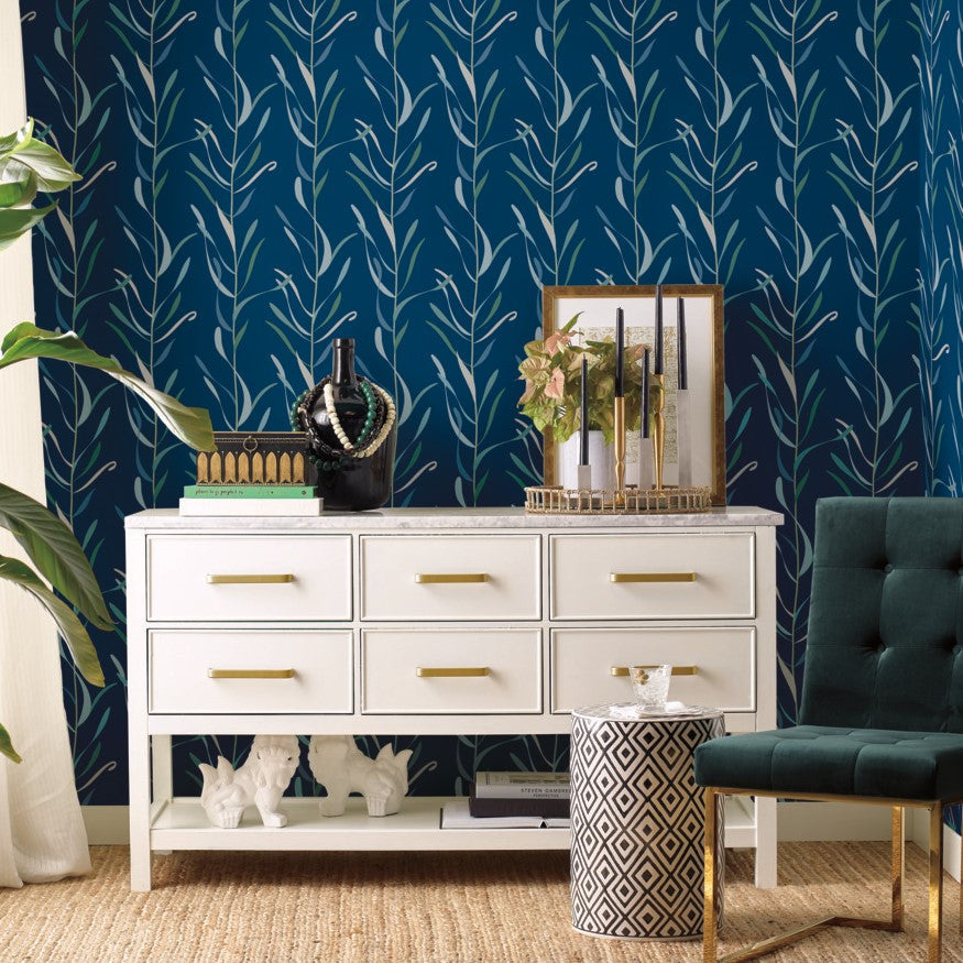 A stylish interior setup featuring a white dresser with gold handles against Chloe Vine River Rock Wallpaper Beige, Grey (60 Sq.Ft.) from York Wallcoverings, accompanied by a dark green armchair and decorative items on the dresser.