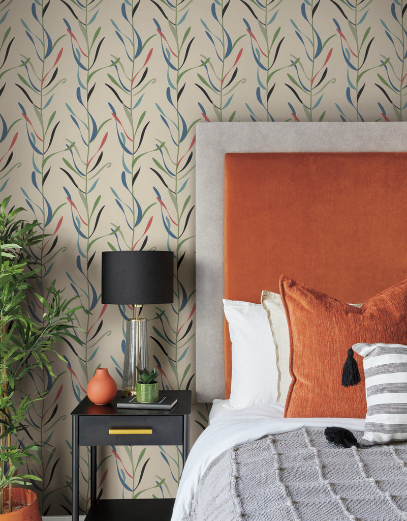 A cozy bedroom corner featuring a bed with a rust-colored headboard, white bedding, and orange pillows, next to a black side table with a lamp, all against a York Wallcoverings Chloe Vine River Rock Wallpaper Beige, Grey (60 Sq.Ft.).