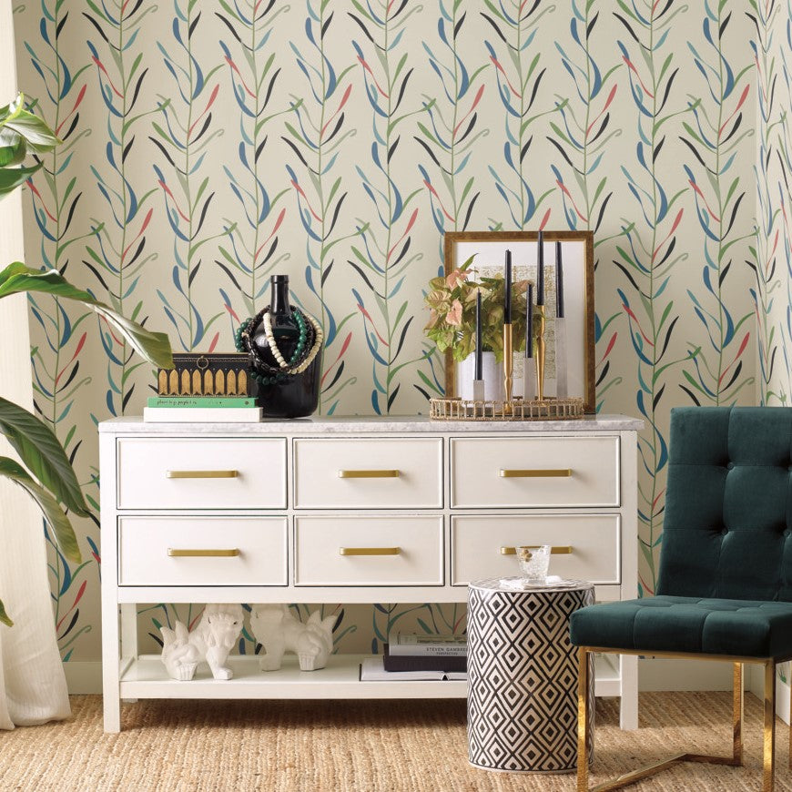 A stylish interior featuring a white dresser with gold handles, adorned with decorative items, set against a vibrant York Wallcoverings Chloe Vine River Rock Wallpaper Beige, Grey (60 Sq.Ft.). A green armchair and a small patterned stool complete the.