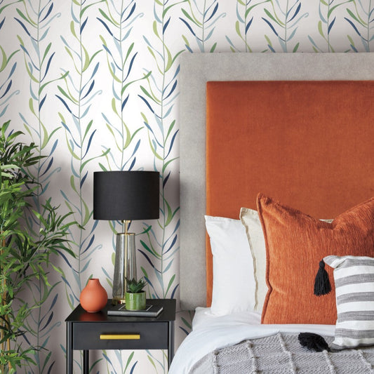 A stylish bedroom features a bed with an orange headboard, white and orange pillows, and a gray blanket. The nightstand beside the bed holds a black and gold lamp, a glass and wooden tray, and decor items. The whimsical design is completed with York Wallcoverings Chloe Vine Peacock Wallpaper Blue, Green (60 Sq.Ft.) in leafy green, blue, and beige.