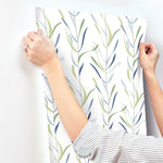 A person wearing a striped shirt is applying York Wallcoverings Chloe Vine Indigo Wallpaper Blue, Green (60 Sq.Ft.) with a whimsical design of green and blue leaves to a white wall. The person's hands are smoothing out the wallpaper, and a ring is visible on their finger.