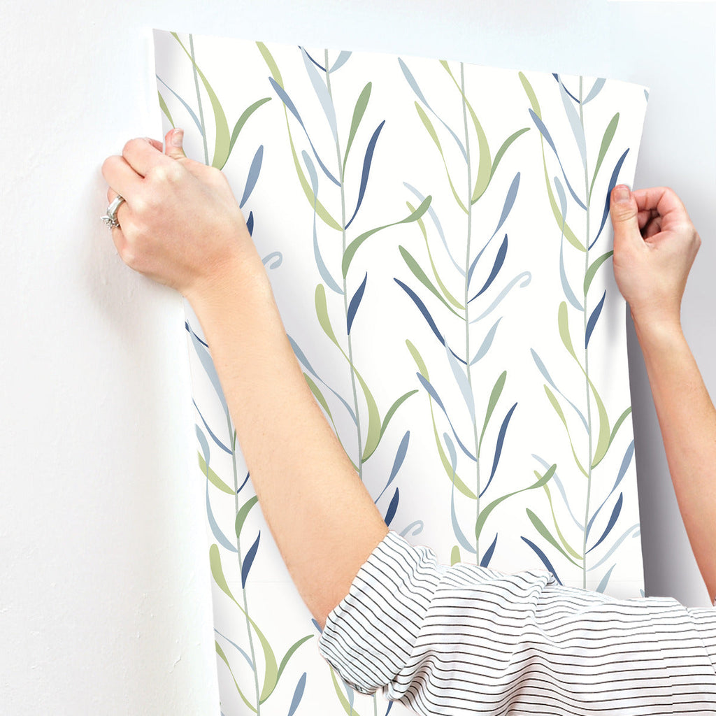 A person hanging a large sheet of Chloe Vine River Rock Wallpaper Beige, Grey (60 Sq.Ft.) with a green and blue leaf pattern by York Wallcoverings on a white wall.