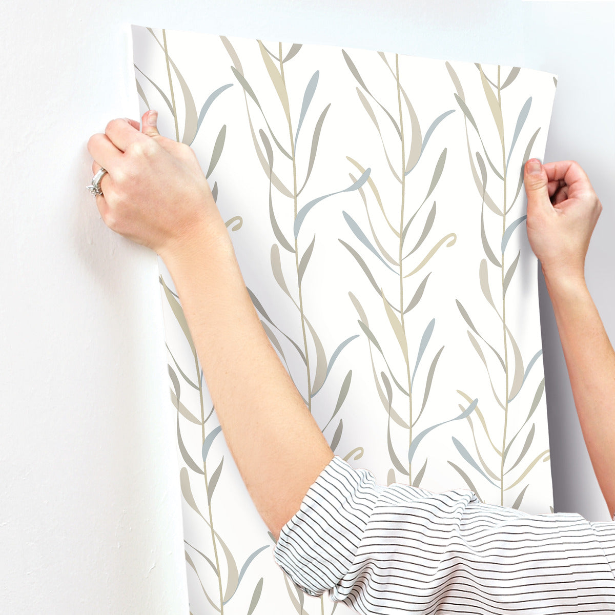 A person in a striped shirt is applying York Wallcoverings Chloe Vine Indigo Wallpaper Blue, Green (60 Sq.Ft.) to a wall. The whimsical design features thin, flowing stems and leaves in muted colors, such as beige and light blue. Their hands are holding the SureStrip wallpaper in place, smoothing it out against the wall.