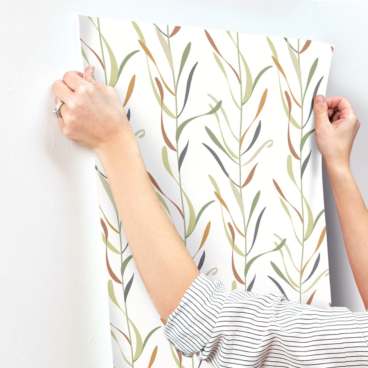 A person is hanging a York Wallcoverings Chloe Vine River Rock Wallpaper Beige, Grey (60 Sq.Ft.) with a delicate green and brown leaf pattern on a white wall; only their arms and hands are visible.