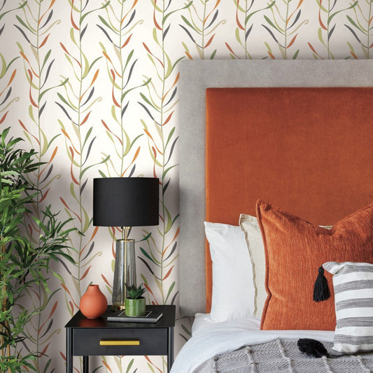 A modern bedroom features a large orange headboard, whimsical leaf-patterned Chloe Vine Sienna Wallpaper Orange, Green (60 Sq.Ft.) by York Wallcoverings, and a gray side table. The table holds a black lamp with a white shade, an orange vase, and a small plant. A gray blanket with tassels is draped over the bed.